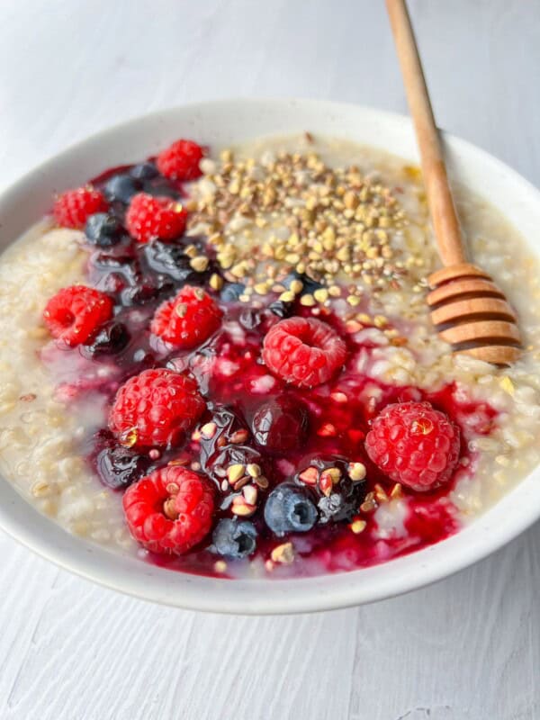 A bowl of oatmeal topped with fresh raspberries, blueberries, a honey drizzle, and a sprinkle of seeds, served with a wooden honey dipper on a light wooden table.