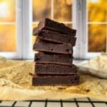 A stack of six chocolate brownies is displayed on parchment paper. The background features a blurred view of a window with autumn foliage visible outside. The scene is warmly lit, creating a cozy atmosphere.