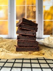 A stack of six chocolate brownies is displayed on parchment paper. The background features a blurred view of a window with autumn foliage visible outside. The scene is warmly lit, creating a cozy atmosphere.