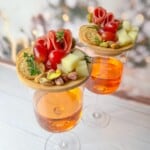 Two glasses of orange-colored drinks topped with small wooden boards featuring appetizers. Each board has salami, cheese cubes, cherry tomatoes, crackers, pistachios, and fresh herbs. Background shows soft-focus holiday decorations.
