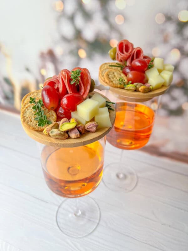 Two glasses of orange-colored drinks topped with small wooden boards featuring appetizers. Each board has salami, cheese cubes, cherry tomatoes, crackers, pistachios, and fresh herbs. Background shows soft-focus holiday decorations.