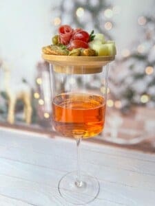A wine glass filled with orange liquid, topped with a wooden board holding cheese cubes, olives, cherry tomatoes, and slices of prosciutto. Blurry, festive lights in the background create a cozy atmosphere.