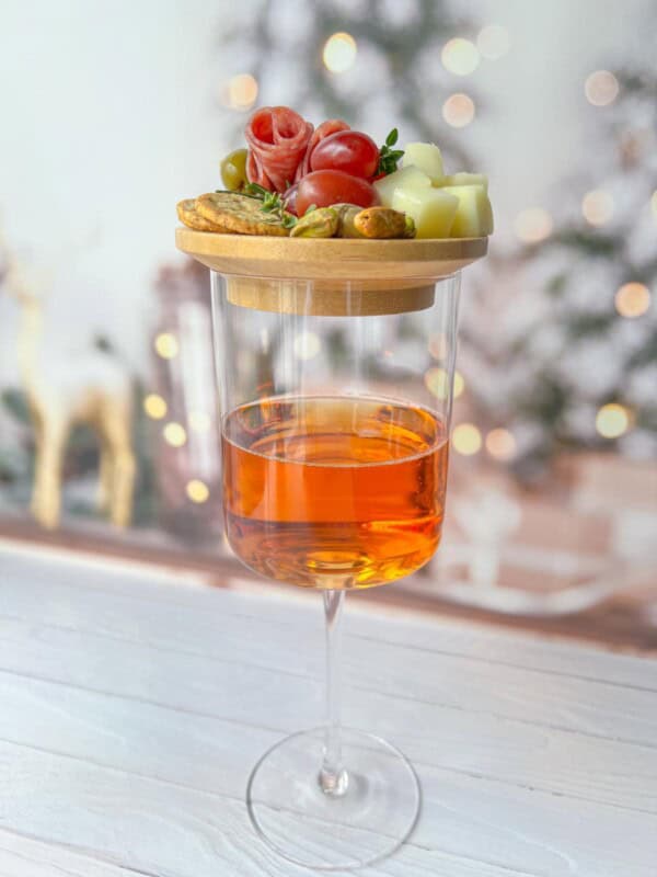 A wine glass filled with orange liquid, topped with a wooden board holding cheese cubes, olives, cherry tomatoes, and slices of prosciutto. Blurry, festive lights in the background create a cozy atmosphere.