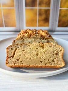 A sliced loaf of moist blender banana bread on a white plate, displayed in front of a window with autumn foliage visible outside. The bread has a golden crust and is topped with chopped nuts.