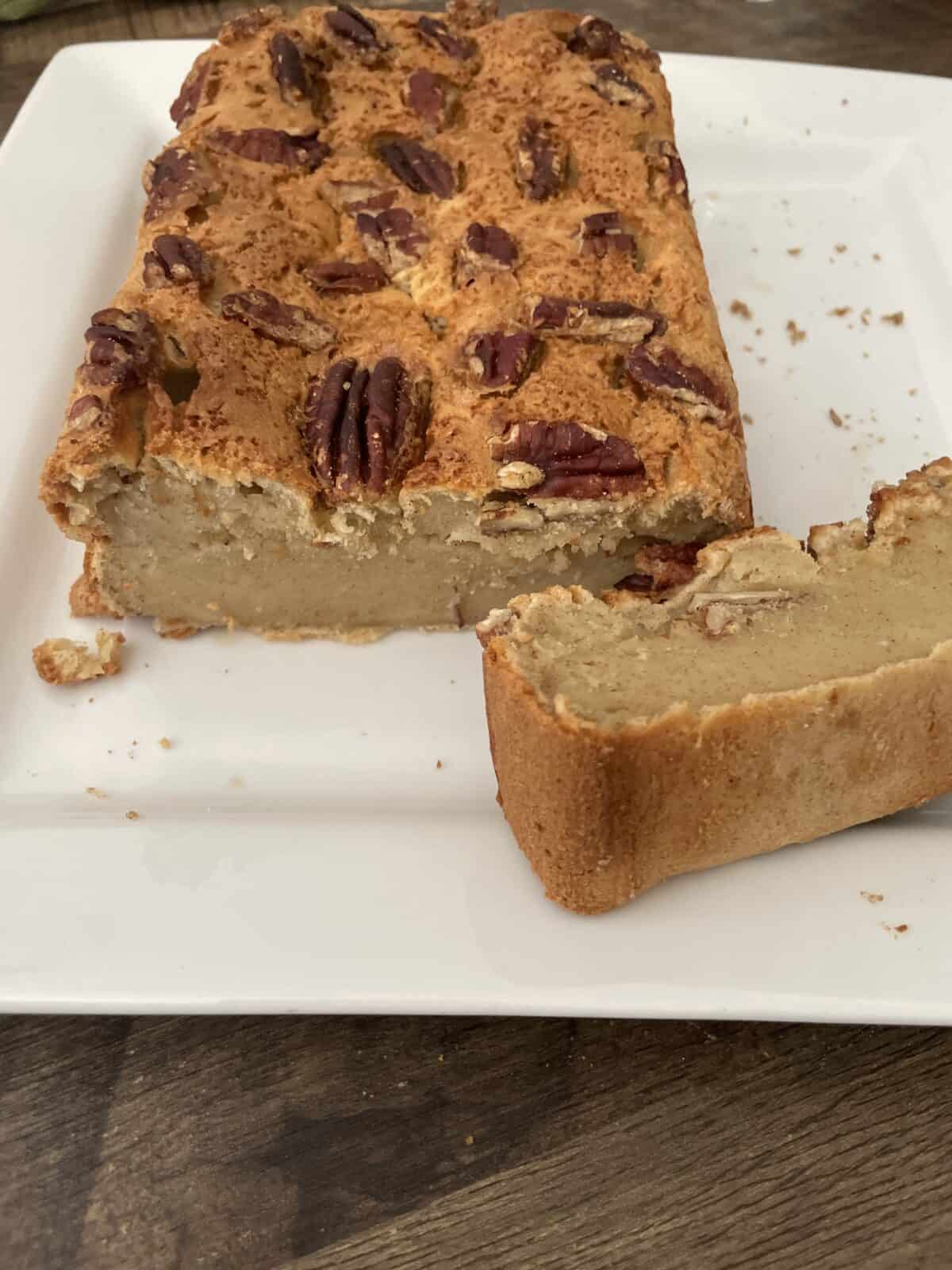 A loaf of cake topped with pecans sits on a white plate, with one thick slice cut and placed in front. The cake has a dense, moist texture and a golden-brown crust.