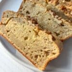 Sliced loaf of banana bread with chopped walnuts on top, displayed on a white plate. The bread is golden brown, showcasing a moist and fluffy texture.