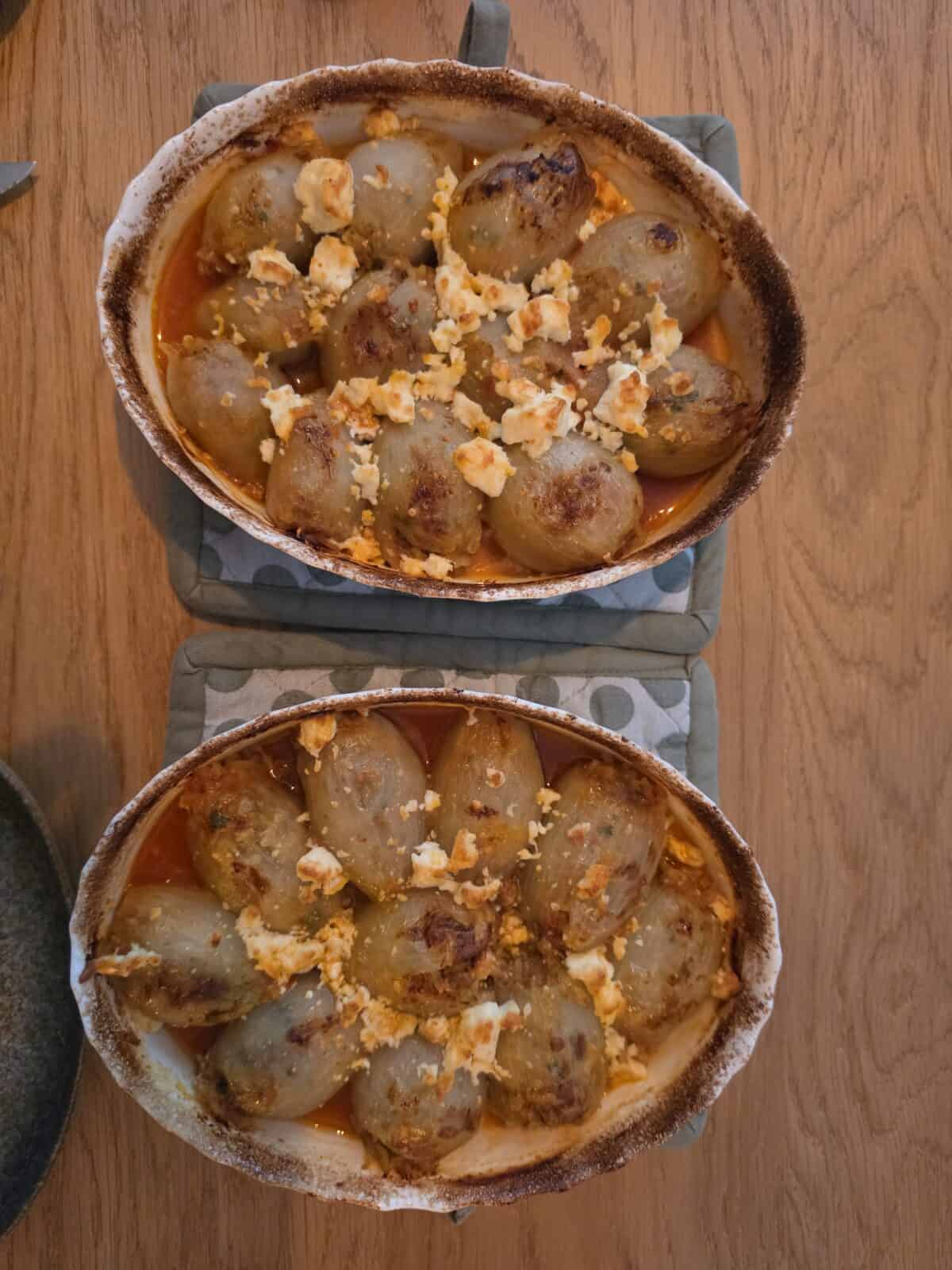 Two oval baking dishes filled with savory Stuffed Onions, topped with crumbled cheese and baked to a golden brown, sit on fabric potholders on a wooden table.