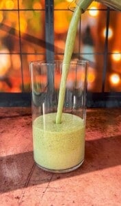 A green smoothie is being poured into a clear glass, set against a warm, blurred background of lights and a window. The glass sits on a wooden surface.