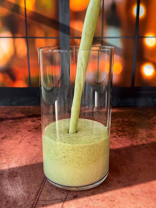 A thick, green smoothie is being poured into a clear glass. The background features a warm, out-of-focus display of glowing lights, creating a cozy ambiance. The scene is set on a wooden surface.