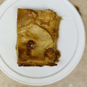 A rectangular slice of apple cake with visible apple slices on top, placed on a white plate against a beige background.