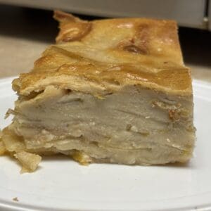 A slice of dense, homemade apple cake with visible layers of apples, sitting on a white plate. The cake has a golden brown, slightly glossy top crust.