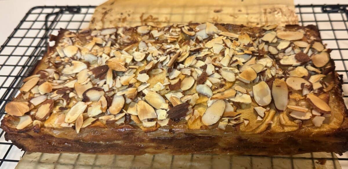 A rectangular Invisible Apple Cake topped with toasted sliced almonds sits on a cooling rack, with a piece of parchment paper partially underneath. The cake has a golden-brown crust.