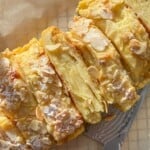 A close-up view of a sliced almond-topped pastry, dusted with powdered sugar. The golden layers of the pastry are visible, and a silver knife lies beside it on a wooden surface.