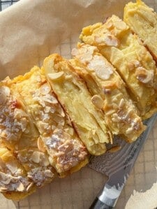 A close-up view of a sliced almond-topped pastry, dusted with powdered sugar. The golden layers of the pastry are visible, and a silver knife lies beside it on a wooden surface.