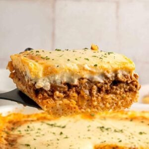 A close-up of a spatula lifting a slice of baked pasta with layers of meat sauce, creamy béchamel, and melted cheese, sprinkled with herbs. The remaining dish is visible in the background.