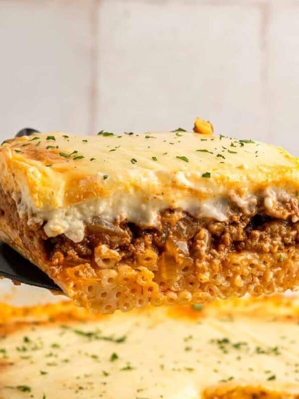 A close-up of a spatula lifting a slice of baked pasta with layers of meat sauce, creamy béchamel, and melted cheese, sprinkled with herbs. The remaining dish is visible in the background.