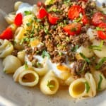 A bowl of pasta with creamy sauce, topped with seasoned ground meat, cherry tomato halves, and fresh parsley. The dish is served in a light-colored bowl with a spoon. Sunlight casts a warm glow over the meal.