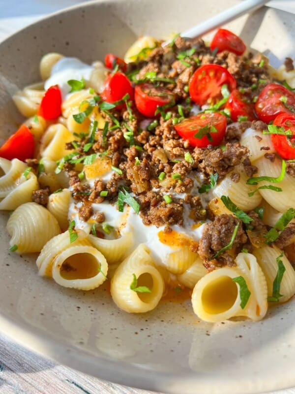 A bowl of pasta with creamy sauce, topped with seasoned ground meat, cherry tomato halves, and fresh parsley. The dish is served in a light-colored bowl with a spoon. Sunlight casts a warm glow over the meal.