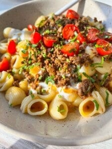 A bowl of pasta shells topped with ground meat, sliced cherry tomatoes, yogurt, and fresh herbs. The dish is garnished with chopped parsley and is bathed in warm sunlight.