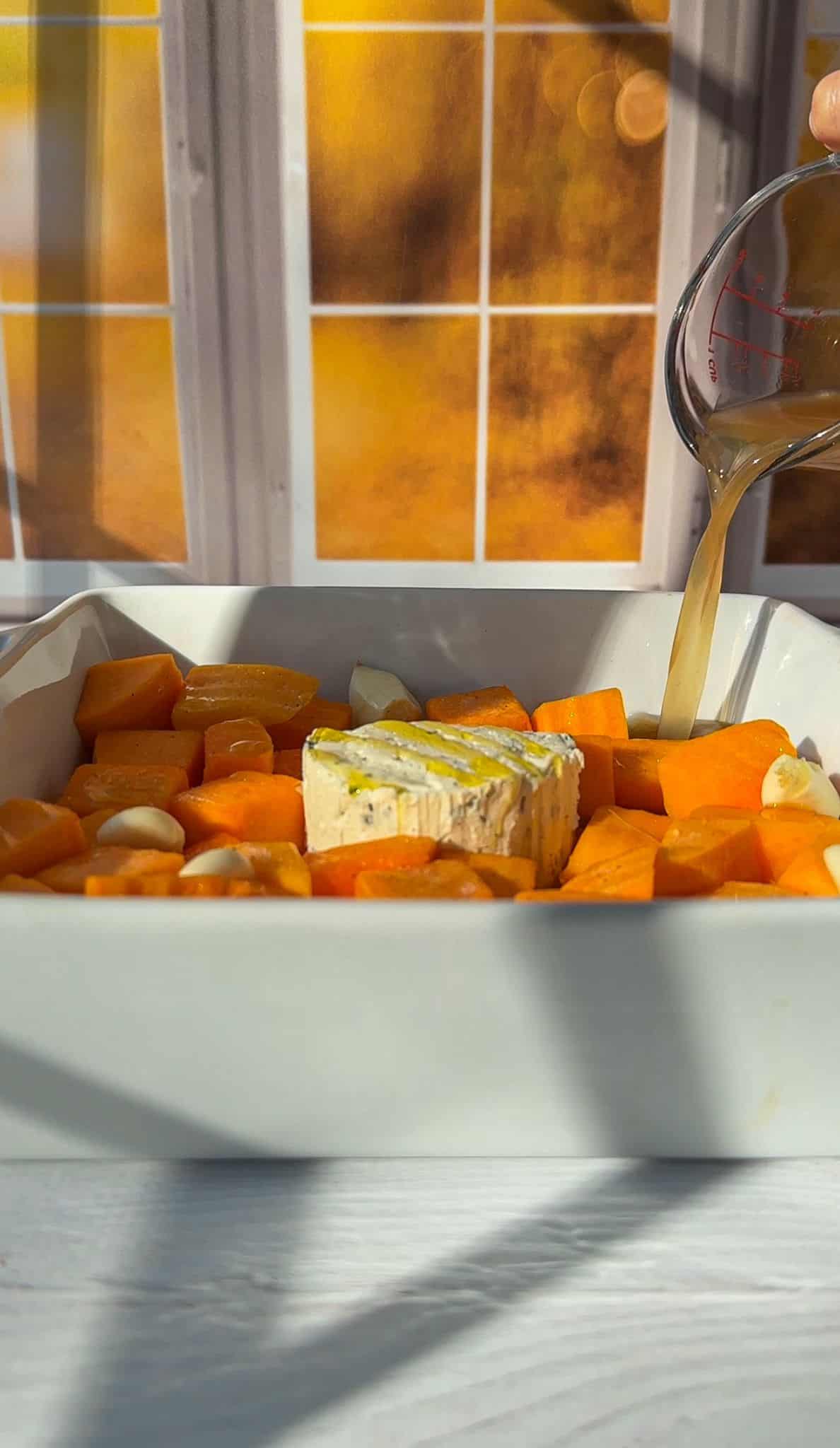 A baking dish filled with cubed sweet potatoes, garlic cloves, and a wheel of brie is shown. A hand is pouring broth from a measuring cup over the ingredients. A sunlit window with a warm orange glow is in the background.