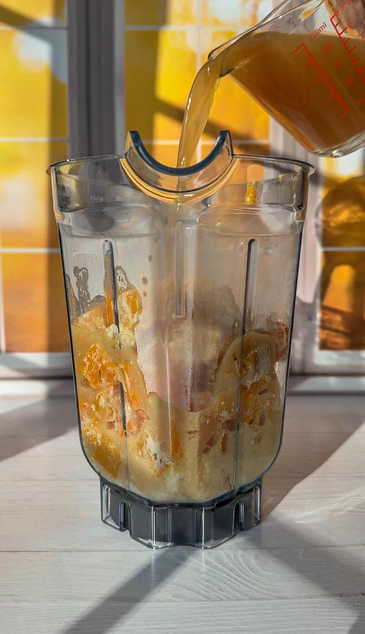 A blender container filled with squash, garlic and Boursin cheese is placed on a white countertop. A measuring cup is pouring a liquid into the blender. Sunlight filters through a window in the background, casting warm shadows. - 5