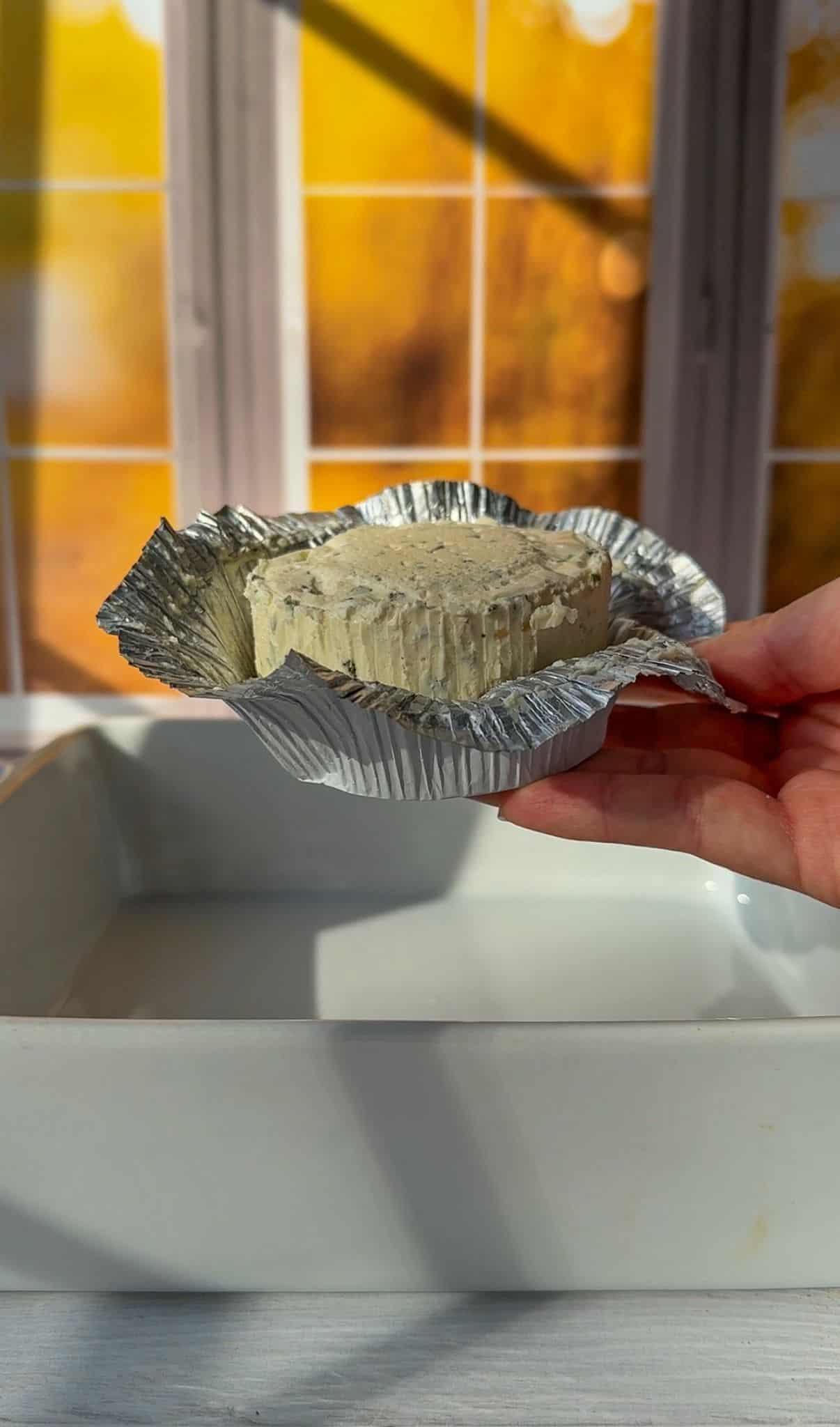 A hand holding an opened foil-wrapped round of cheese over a white dish. Warm sunlight streams through a window with a grid pattern in the background. - 4