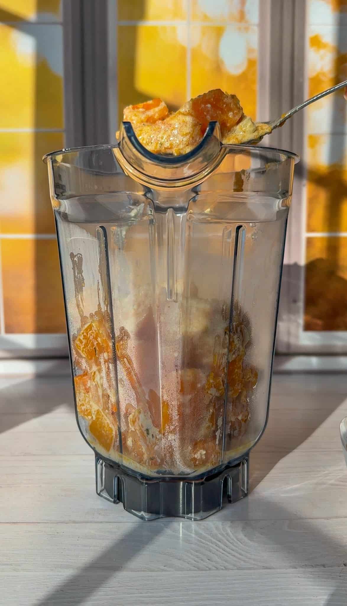 A blender jar filled with sliced bananas and oranges is placed on a kitchen counter. A spoon is lifting some of the fruit mixture out. Sunlight filters through a window behind, casting warm yellow and orange hues.