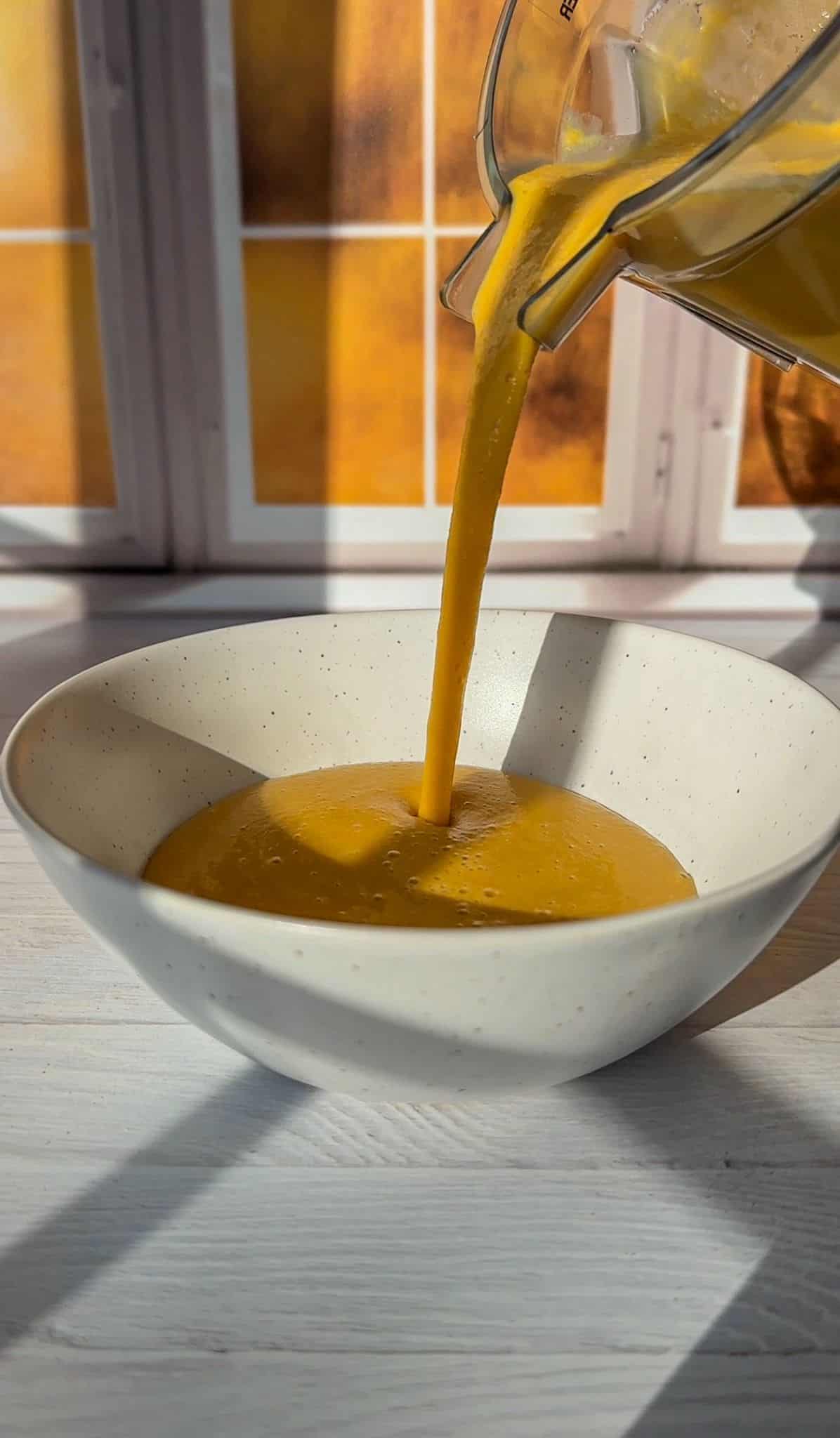 Smooth butternut squash soup being poured into a white speckled ceramic bowl, with a backdrop of sunlit windows.