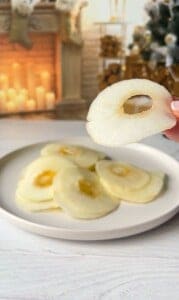 A hand holding a thinly sliced apple in front of a plate with more apple slices. In the background, there's a lit fireplace and a decorated Christmas tree.