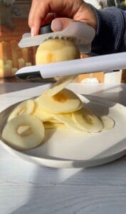 A hand uses a slicer to cut an apple into thin, uniform rings on a white plate. Sunlight casts shadows on the white wooden surface, and several sliced apple rings are neatly piled.