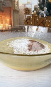 A glass bowl filled with cake batter and topped with flour and cocoa sits on a white kitchen counter. In the background, a decorated Christmas tree and a lit fireplace are visible, creating a cozy holiday atmosphere.