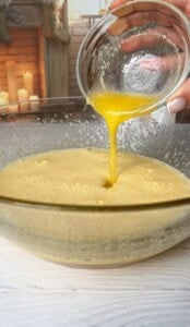 A close-up of a hand pouring melted butter from a small glass bowl into a larger bowl containing a batter mixture. A warm, cozy background with lit candles and a wooden fireplace is visible.