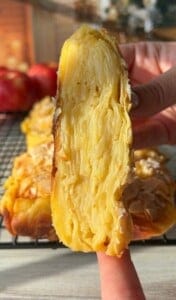Close-up of a hand holding a slice of layered apple dessert, showcasing the intricate layers within. The dessert has a golden crust, and sliced almonds are sprinkled on top. A cooling rack and red apples are visible in the blurred background.