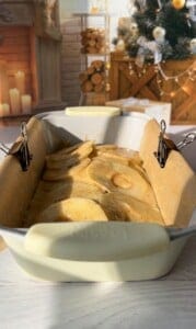 A loaf pan lined with parchment paper and secured with binder clips is filled with batter and apple slices. The background shows a cozy room with a decorated Christmas tree, a fireplace, stacked logs, and lit candles.