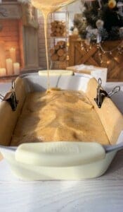 A creamy batter is being poured into a loaf pan lined with parchment paper, secured with binder clips. The background features a cozy fireplace and a decorated Christmas tree, creating a warm holiday atmosphere.