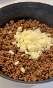 A black skillet filled with cooked ground beef topped with chopped white onions. The mixture is being prepared on a stovetop, with the onions yet to be stirred into the beef.
