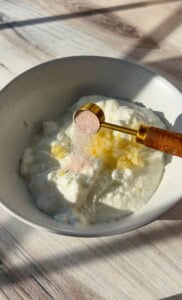 A bowl of white yogurt with finely chopped garlic. A golden measuring spoon holds pink salt, ready to be added to the mixture. The bowl sits on a light wooden surface with soft shadows.