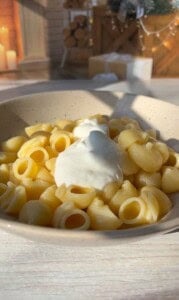Bowl of pasta shells topped with a dollop of cream sauce. The background shows a cozy room with a pile of logs, candles, and festive decorations. Sunlight streams in, casting shadows on the table.