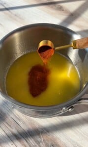 A tablespoon of red spice is being poured into a saucepan containing melted butter, set on a light-colored wooden surface. The sunlight casts gentle shadows across the scene.