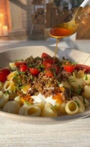 A bowl of pasta with chopped tomatoes, ground meat, and sprinkled herbs. A spoon drizzles a golden sauce over the dish, enhancing its appeal. The background includes candles and wooden decor, adding a cozy atmosphere.