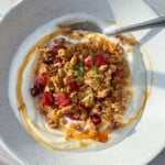 A bowl of creamy yogurt topped with granola, dried cranberries, and seeds, drizzled with honey, garnished with fresh mint. A spoon rests in the bowl, set on a light wood surface with natural sunlight casting shadows.