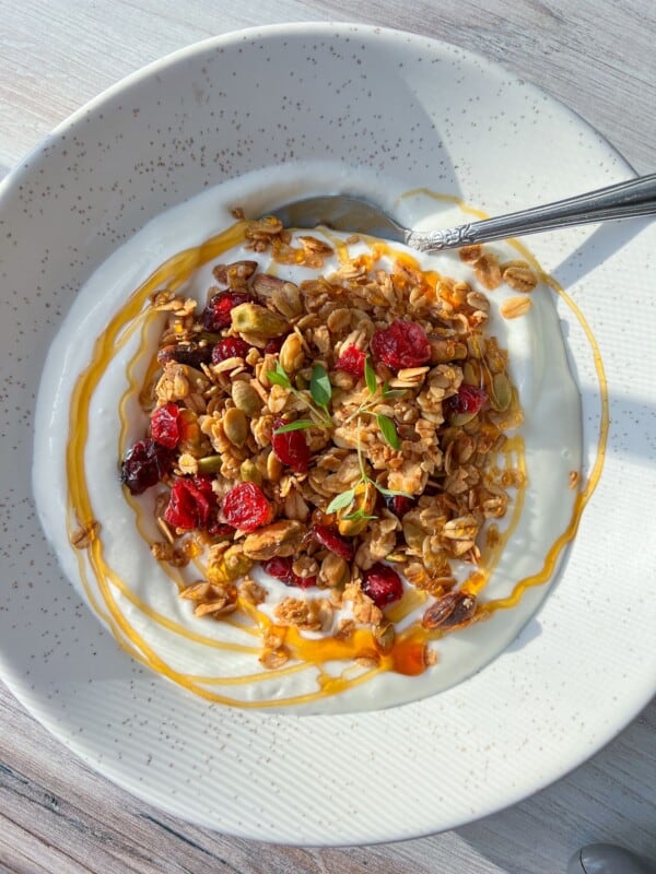 A bowl of creamy yogurt topped with granola, dried cranberries, and seeds, drizzled with honey, garnished with fresh mint. A spoon rests in the bowl, set on a light wood surface with natural sunlight casting shadows.
