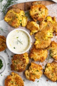 Crispy smashed potatoes garnished with herbs are spread on parchment paper. A small bowl of creamy dip, garnished with dill, is placed to the side. Fresh dill and chopped herbs are scattered around, enhancing the rustic presentation.