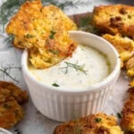 A piece of fried corn fritter is dipped into a small white ramekin filled with creamy herb sauce. Additional fritters are scattered around on a light surface, garnished with fresh dill leaves.
