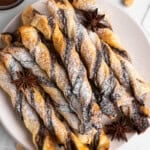 A plate of twisted puff pastry sticks with chocolate, dusted with powdered sugar. Star anise and hazelnuts are scattered around the plate. A small bowl of chocolate sauce is visible in the corner.