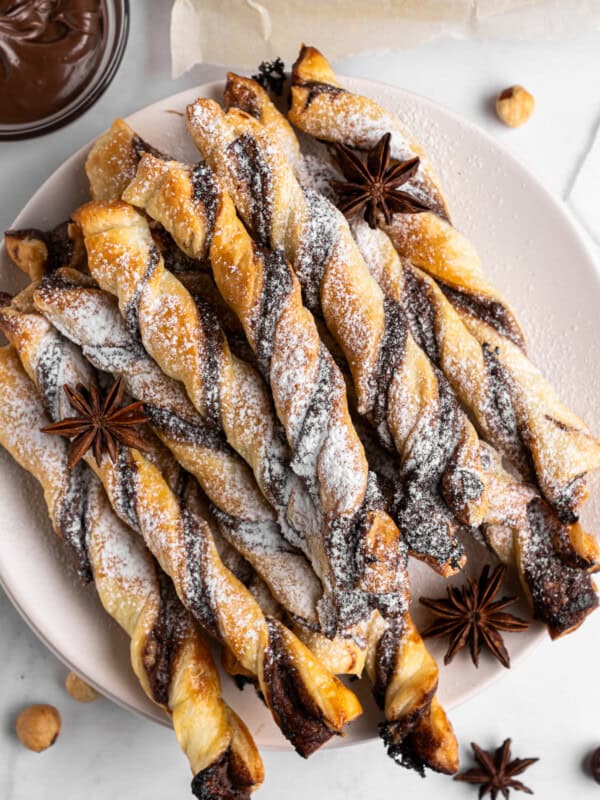 A plate of twisted puff pastry sticks with chocolate, dusted with powdered sugar. Star anise and hazelnuts are scattered around the plate. A small bowl of chocolate sauce is visible in the corner.