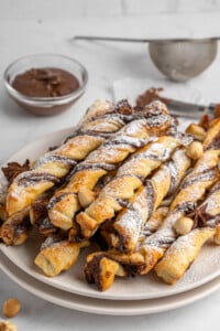 A plate of twisted pastry sticks dusted with powdered sugar. The pastries are golden brown with a chocolate filling. A small bowl of chocolate spread and a sieve are in the background, along with a few scattered nuts.