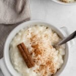 A bowl of rice pudding topped with cinnamon powder and a cinnamon stick, with a spoon on the side. A second bowl is partially visible. A beige cloth napkin is beside the bowl, on a white marble surface.