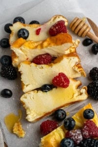 Slices of cheesecake topped with blackberries, raspberries, and blueberries on parchment paper. A honey dipper with honey is nearby, and some honey is drizzled over the cheesecake.