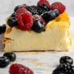 Slice of creamy cheesecake topped with fresh raspberries, blueberries, and blackberries on a white marble surface. More berries and a drizzle of honey are around the cheesecake, with a blurred white brick background.